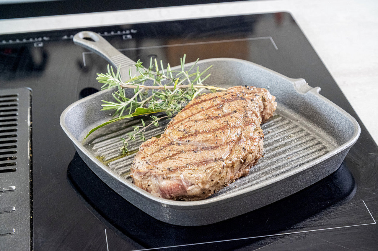 Čuguna grila panna ar steiku uz plīts virsmas, piemērota cepšanai, grilēšanai un gatavošanai virtuvē