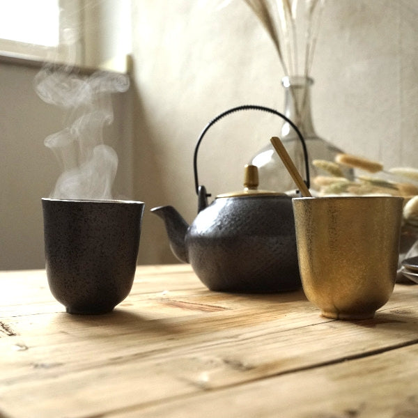 Melna tējkanna ar tvaiku un krūzes uz koka galda, piemērotas tējas pagatavošanai un pasniegšanai mājās